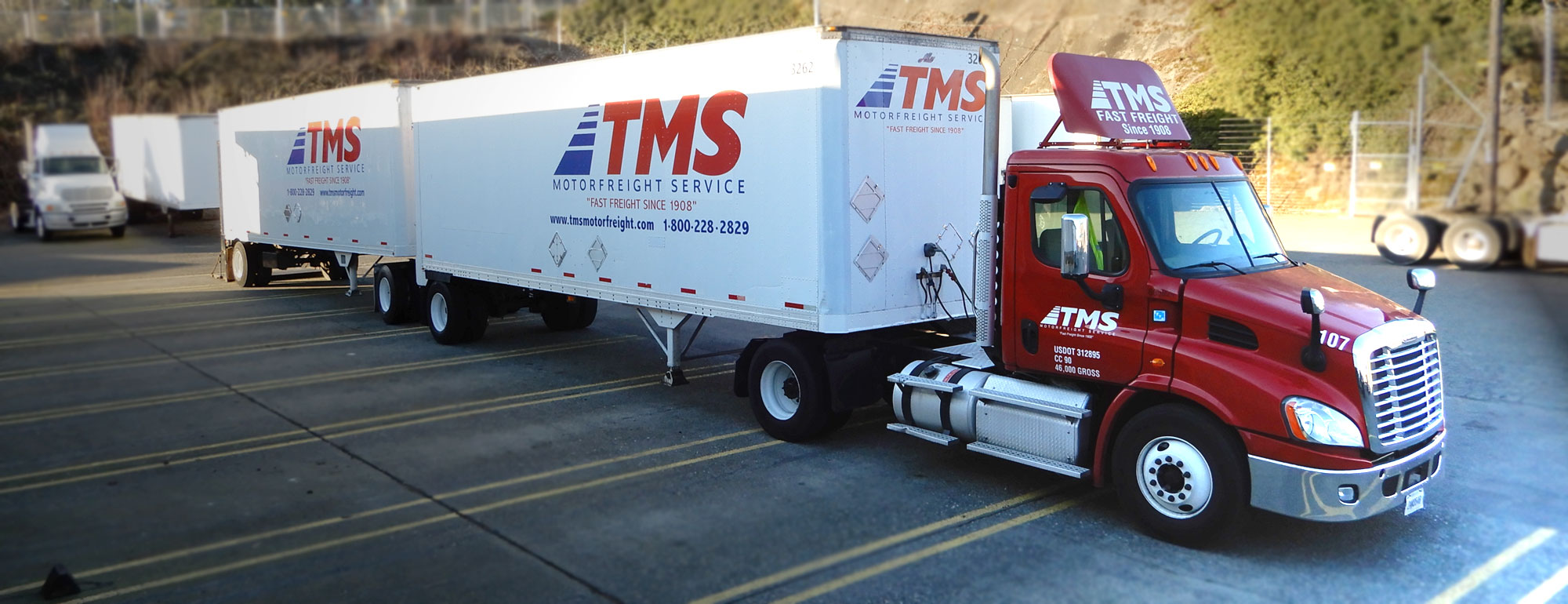 Red TMS semi tractor hooked up to two transport van trailers in a parking lot